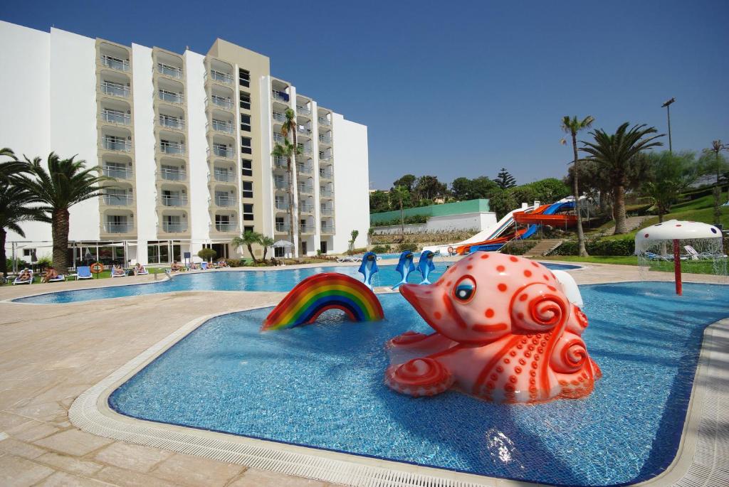 Vasaras brīvdienu komplekss ar peldbaseinu, ko rotā krāsains gumijas astoņkājis un varavīksne, apkārt palmām un viesnīcas ēkai. Ideāla vieta atpūtai un izklaidei.