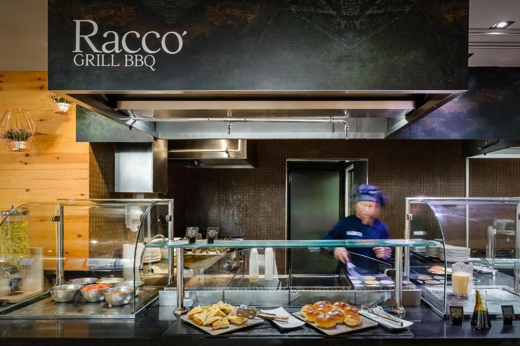 Grila restorāna Racco BBQ virtuves skats ar šefpavāru, kas strādā pie grila, un dažādiem ēdieniem uz galda.