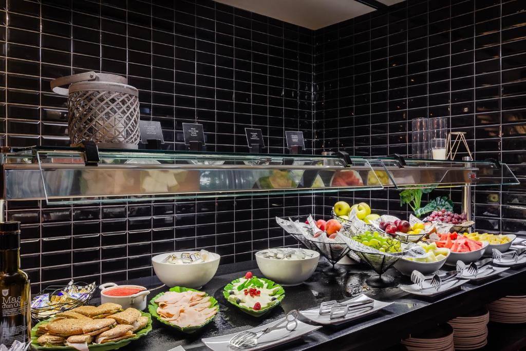 Bagātīgs bufetes galds ar dažādiem augļiem, dārzeņiem un gaļas izstrādājumiem, uz melnas flīžu fona. Iekļauti arī saldumi un dažādas mērces, kas pieejamas viesu izvēlei.