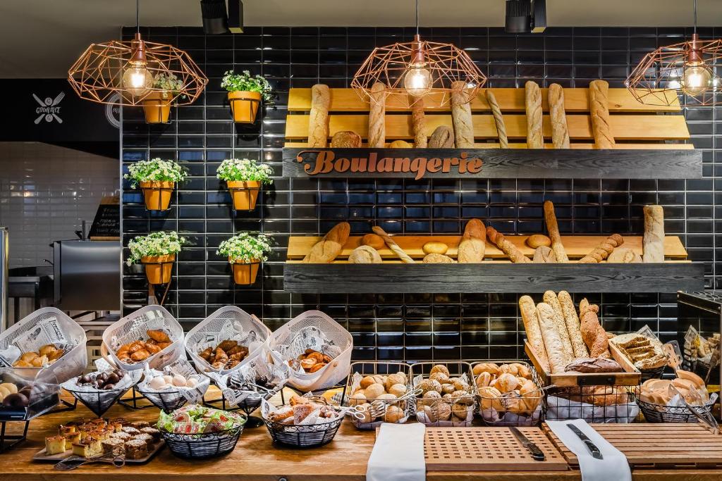 Maizes veikals ar dažādām maizes izstrādājumiem un konditorejas izstrādājumiem, kas sakārtoti grozos uz koka galda. Fons ar melno flīzēm un dekoratīvām augiem.