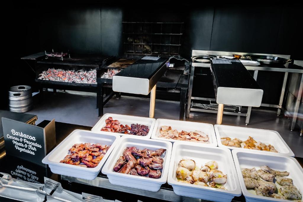 Grila stends ar dažādām gaļas un zivju porcijām, kas izkārtotas baltās traukos. Aiz traukiem redzama ogļu grila zona, kur tiek gatavoti ēdieni.