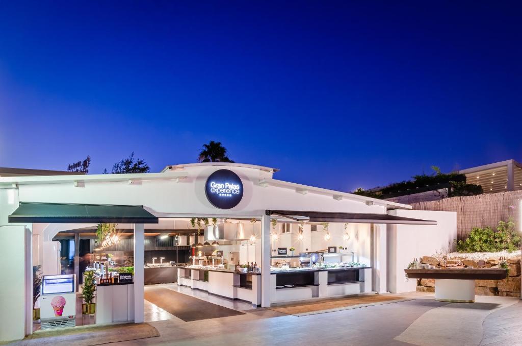 Moderns restorāns "Gran Palas experiencias" ar atvērtu terasi, kas apgaismota siltā gaismā. Fons ir tumši zils debesu krāsojums vakara stundā.