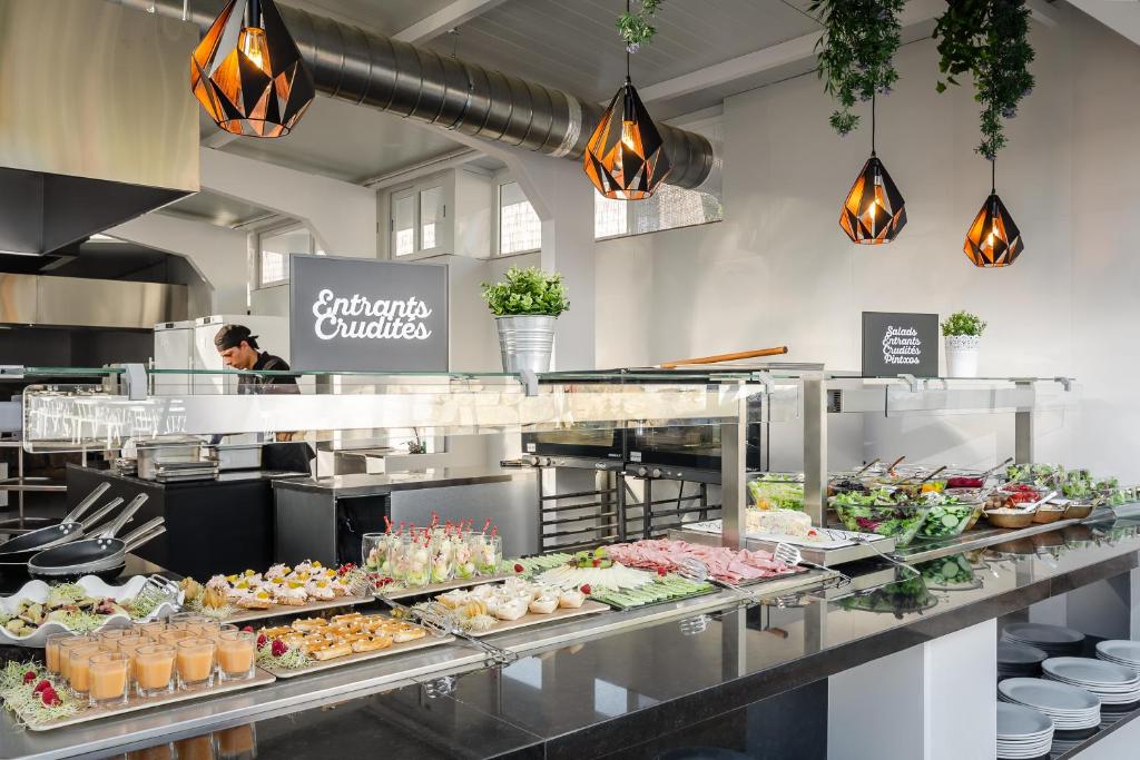 Eleganta pašapkalpošanās zona ar dažādiem uzkodu un salātu piedāvājumiem, ieskaitot dārzeņu šķīvjus, desiņas un desertus. Interjers ir gaišs un mūsdienīgs ar stilīgām lampām un zaļiem augiem.
