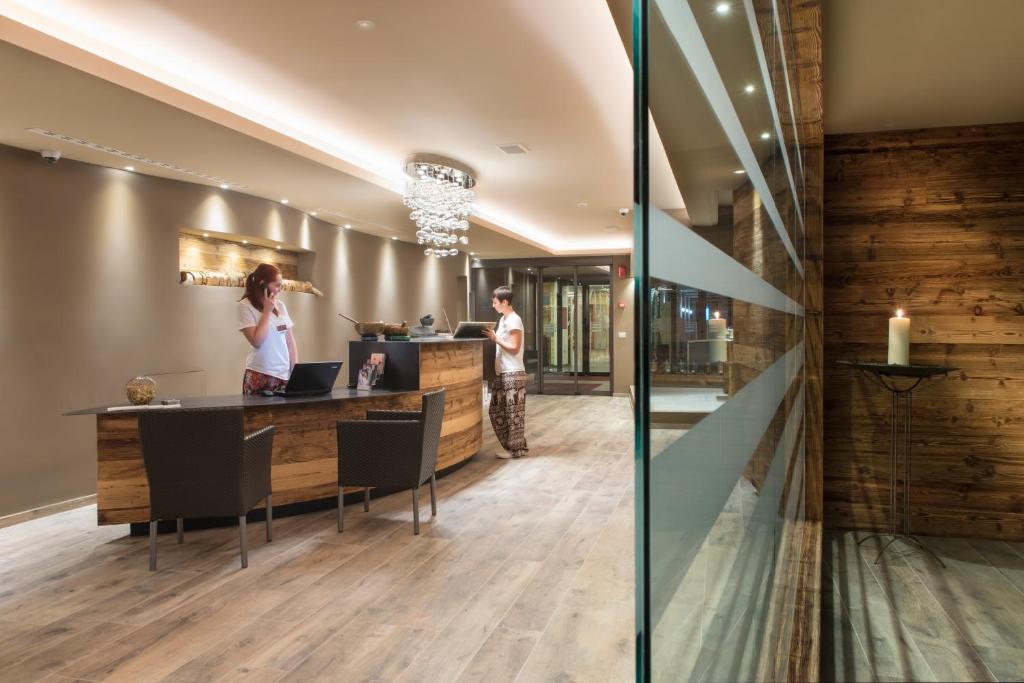 Mūsdienīga viesnīcas reģistratūra ar koka akcentiem un elegantu apgaismojumu, kur divi darbinieki apkalpo viesus. Interjers ir siltā tonī, radot patīkamu atmosfēru.