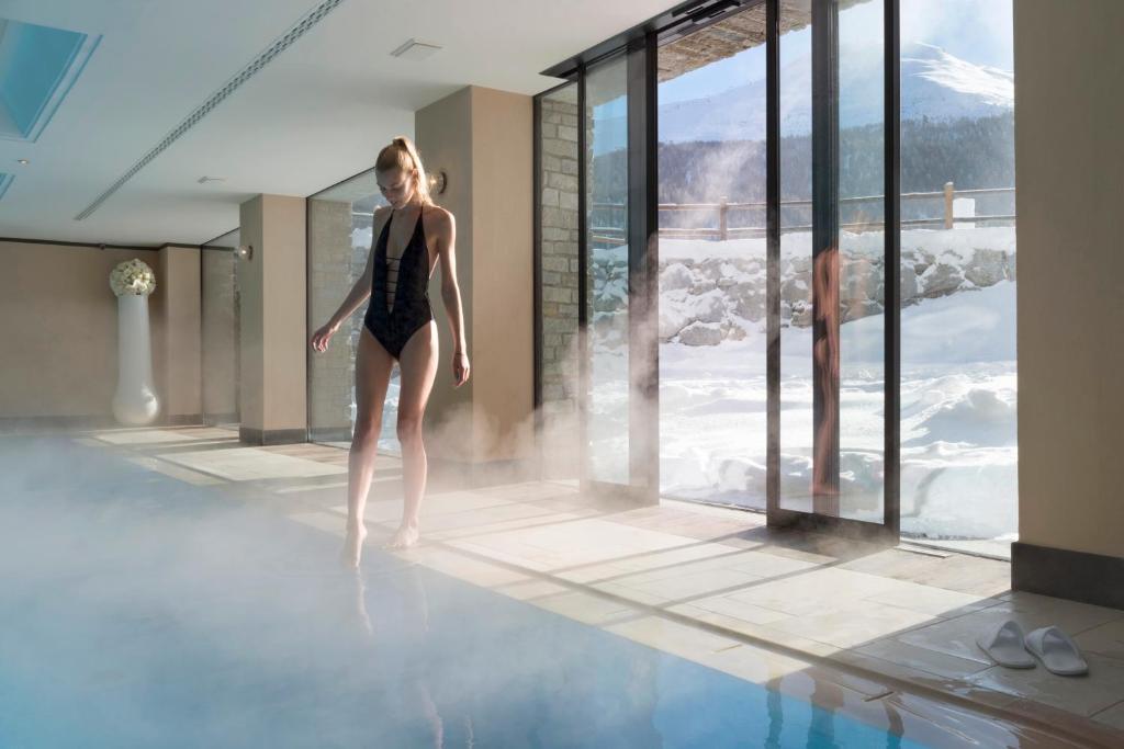 Sieviete melnā peldkostīmā, stāvot pie baseina ar tvaiku, viesnīcas iekštelpās, caur stikla durvīm redzama sniegota ainava.