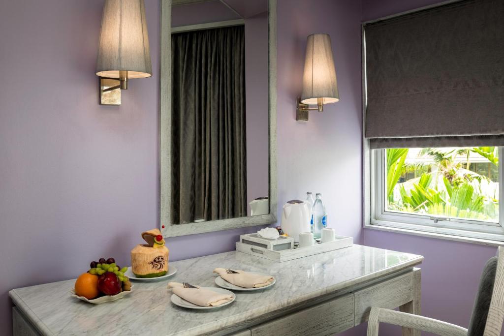 Eleganta viesnīcas istaba ar gaiši violetām sienām, marmora galdu un logu, caur kuru redzami zaļumi. Uz galda ir augļu šķīvis, kūka un dzērieni.