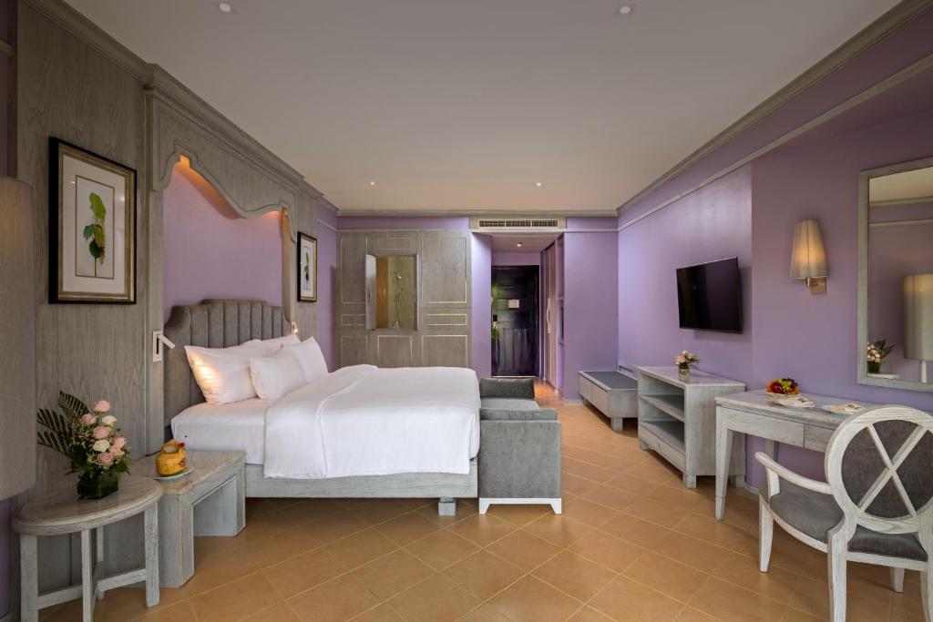 Mūsdienīga viesnīcas istaba ar pelēku gultu un violetām sienām, dekorēta ar ziediem un mākslas darbiem. Istabā ir televizors un ērtas mēbeles.