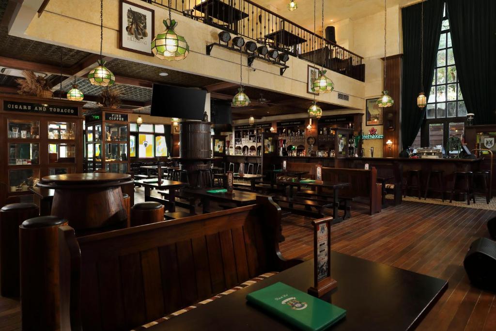 Viesmīlīga krodziņa iekšskats ar koka mēbelēm un zaļiem apgaismojuma elementiem. Gaisma ienāk caur lielajiem logiem, radot siltu atmosfēru.