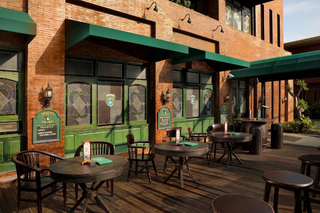 Ārtelpa ar koka galdiem un krēsliem, kas izvietoti uz terases pie restorāna ar sarkanu ķieģeļu sienām un zaļām logu rāmēm. Redzami arī dekoratīvi elementi un ēdamzīmes uz galdiem.