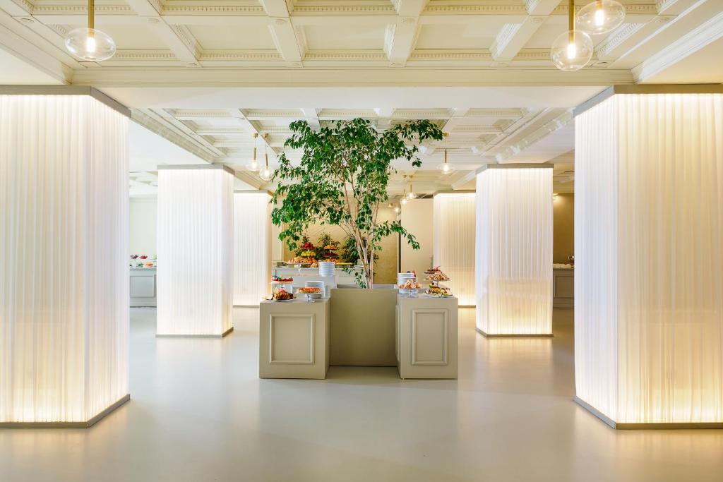 Mūsdienīga kafejnīca ar elegantiem, apgaismotiem stabiem un zaļo augu centrā. Interjers ir gaišs un minimālistisks, radot patīkamu atmosfēru.