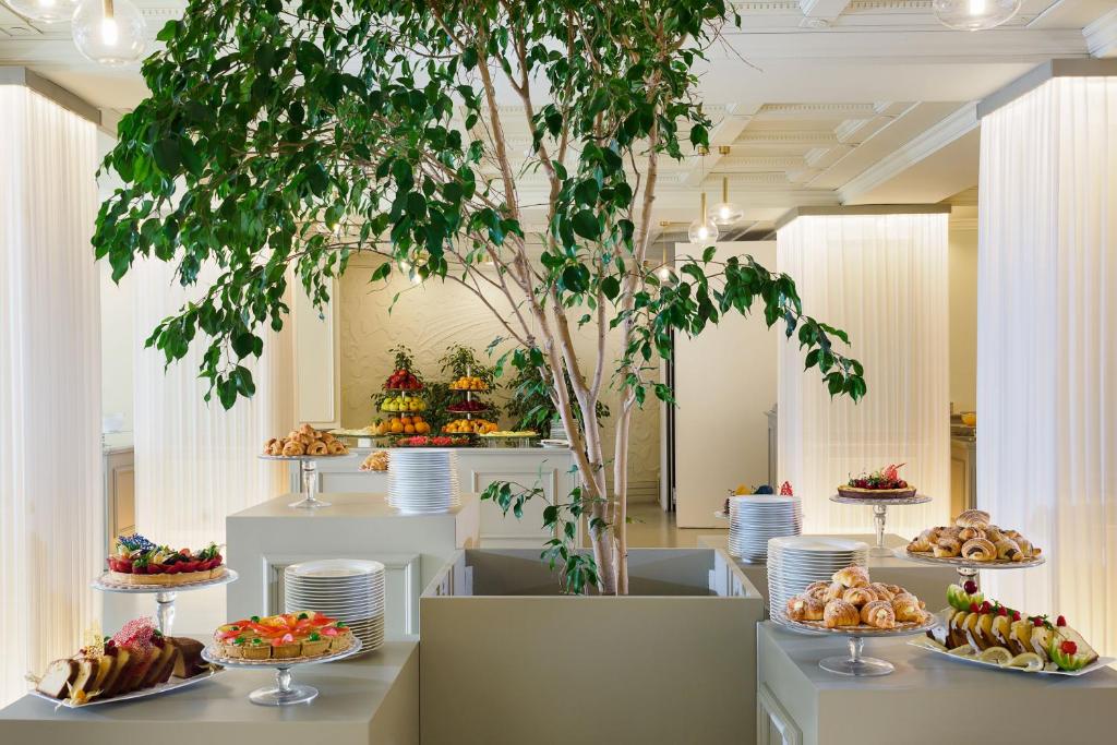 Bagātīgs bufetes galds ar augļiem un konditorejas izstrādājumiem, centrā esošs liels koks, kas piešķir telpai dzīvīgumu. Mūsdienīga un eleganta atmosfēra.