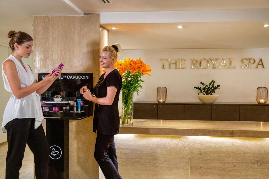 Spa centrā divas sievietes apspriež kosmētikas produktus pie skaisti noformētas uzņemšanas zonas. Fons ir elegants un relaksējošs, ar ziediem un mūsdienīgu dekori.