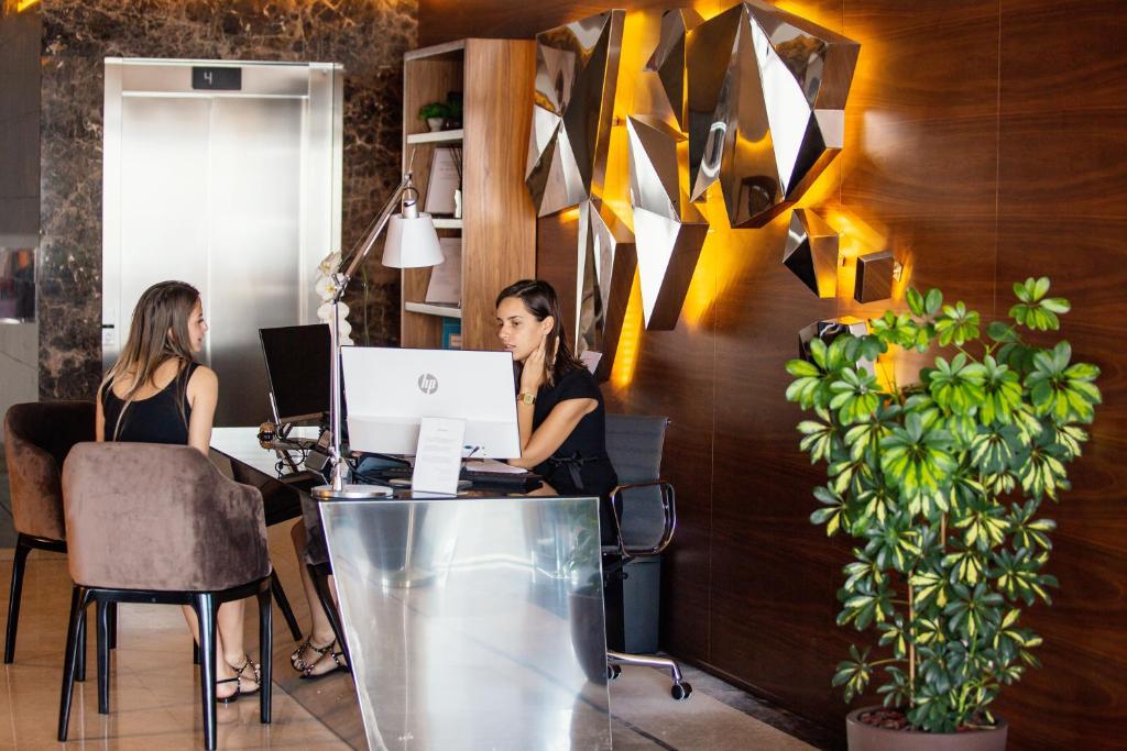 Mūsdienīgs viesnīcas reģistratūras skats, kur divas sievietes sarunājas pie datora. Interjerā redzams stilīgs dekoratīvs elements un zaļa auga klātbūtne.