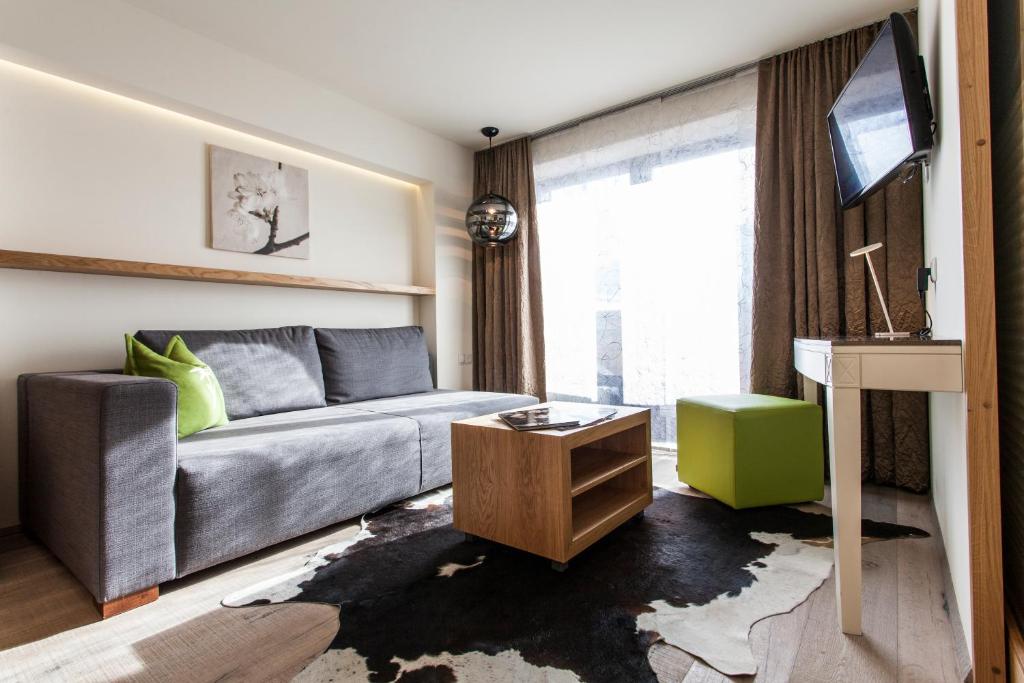 Mūsdienīgs viesistabas interjers ar pelēku dīvānu, koka galdu, zaļu ottomānu un televizoru. Telpa ir gaiša, ar logiem un dabiskiem materiāliem, kas rada komfortablu atmosfēru.