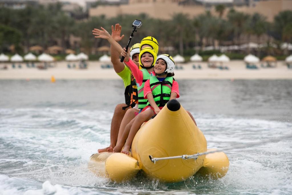 Divi cilvēki, apģērbti glābšanas vestēs, jautri brauc ar banāna laivu ūdenī, ar aizgādni un prieku uz sejas, fonā redzama pludmale un palmām.