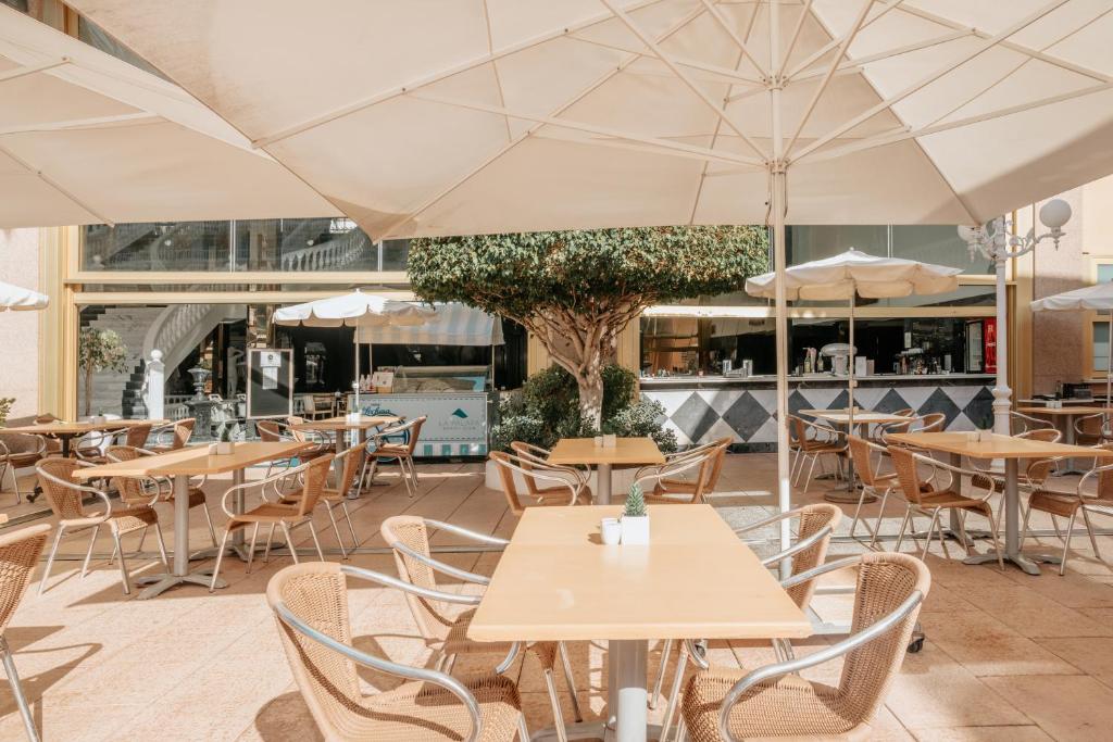 Kafejnīca ar āra terasi, kurā redzami koka galdi un krēsli, kā arī lielas saules parasoles, kas nodrošina ēnu. Aiz terases ir bārs ar kafijas aparātiem.