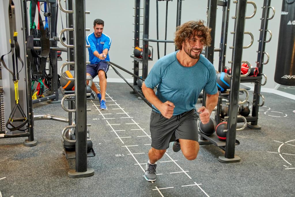 Divi vīrieši sporta zālē intensīvi trenējas, viens skrienot un otrs vilkšanas vingrinājumā. Apkārt ir dažādi trenažieri un sporta inventārs.