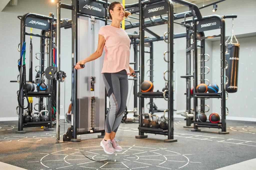 Sieviete lēkā ar lecamauklu modernā fitnesa zālē, apņemta ar sporta aprīkojumu, piemēram, svaru bumbām un trenažieriem.