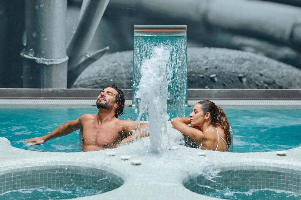 Divi cilvēki relaksējas spa baseinā, izbaudot ūdens strūklu un atpūtu. Aiz viņiem redzama stilīga interjera fona dizains.