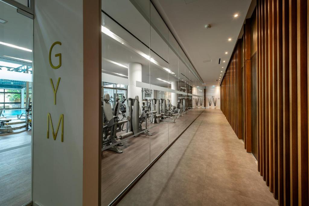 Mūsdienīga sporta zāles ieeja ar stikla sieniņu un koka apdari, kas rada elegantu un atvērtu atmosfēru. Redzami sporta aprīkojumi un labi apgaismota telpa.