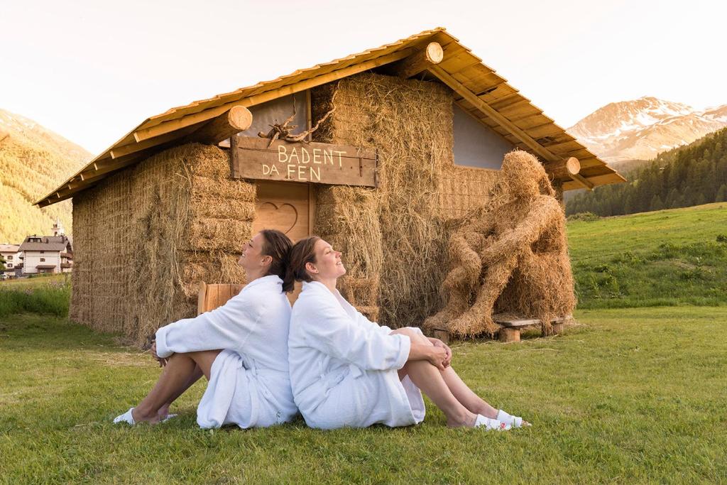 Divas sievietes baltos halātos sēž uz zāliena pie salmu mājas ar zīmējumu "Baden da Fen", kam blakus atrodas salmu skulptūra.