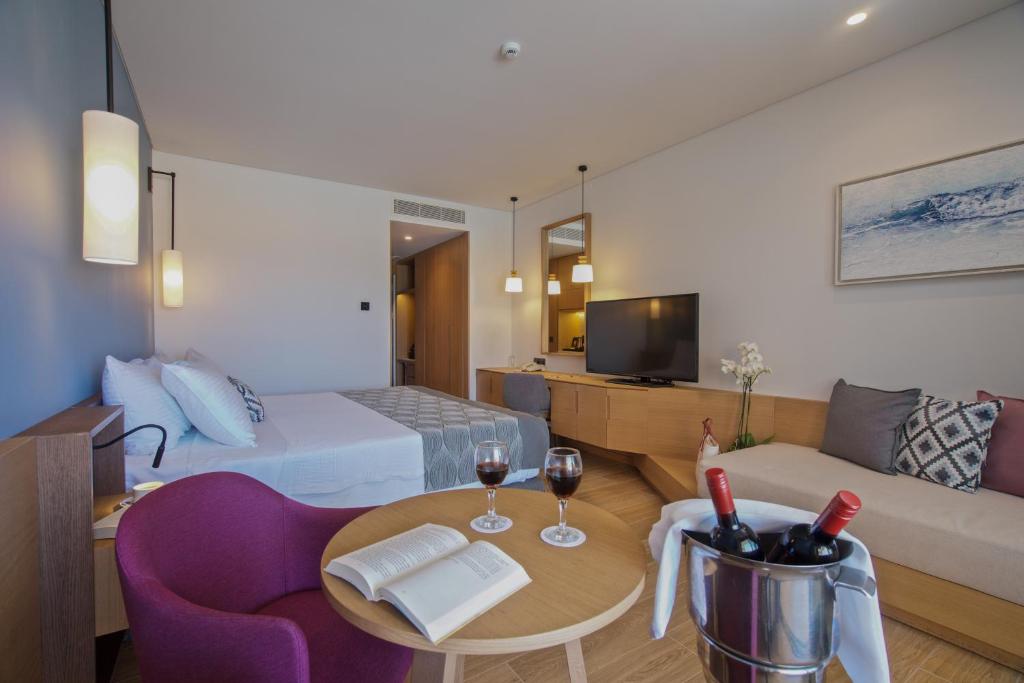 Mūsdienīgs viesnīcas numurs ar ērtu gultu, viesistabu un galdu, kurā ir sarkans krēsls, grāmata un vīna pudeļu grozs. Interjers ir gaišs un elegants, radot relaksējošu atmosfēru.