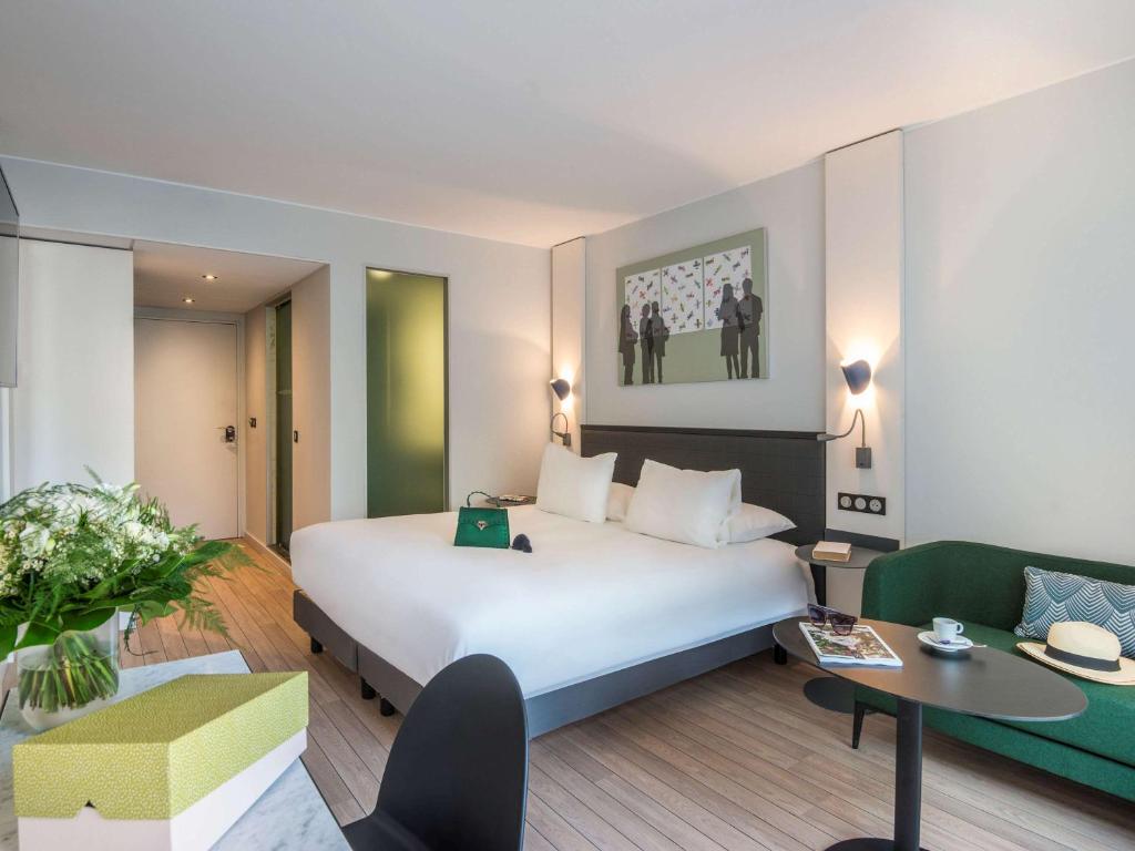 Mūsdienīgs viesnīcas numurs ar plašu gultu, zaļo dīvānu un elegantu galdu. Interjers ir gaišs un stilīgs, ar ziedu kompozīciju un mākslas darbu uz sienas.