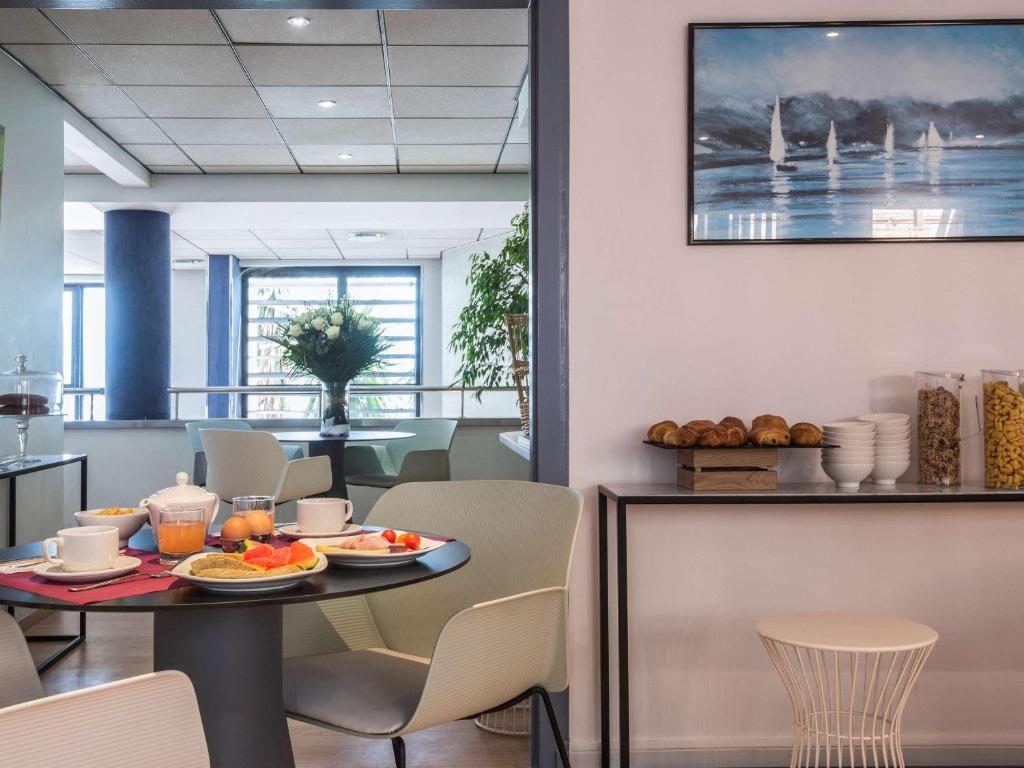 Mūsdienīga brokastu zona ar elegantiem galdiem, dažādiem ēdieniem un dekoratīvām augu kompozīcijām. Gaismas piepildīta telpa ar mākslas darbu pie sienas.