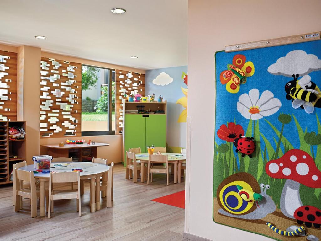 Krāsaina bērnudārza telpa ar galdiem un krēsliem, dekorēta ar košiem ziediem un kukaiņiem uz paklāja. Logi ļauj ienākt dabiskajai gaismai, radot siltu un aicinošu atmosfēru.