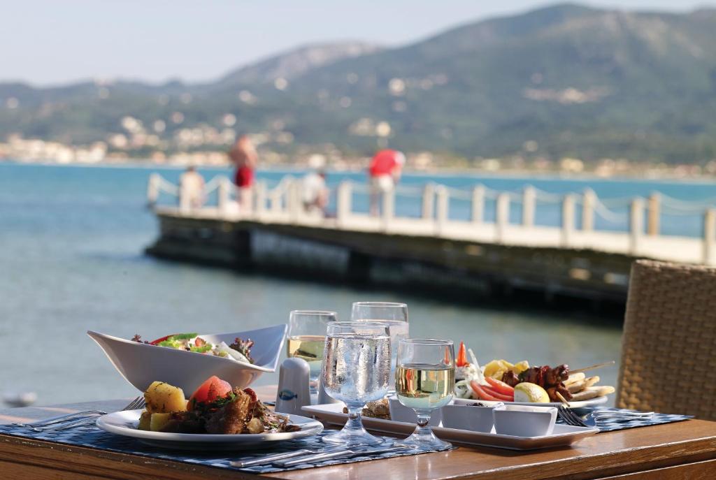 Jūras skats ar viesējo restorānu, kur uz galda ir pasniegti gardi ēdieni un dzērieni, fons ar cilvēkiem, kas pastaigājas pa piestātni.