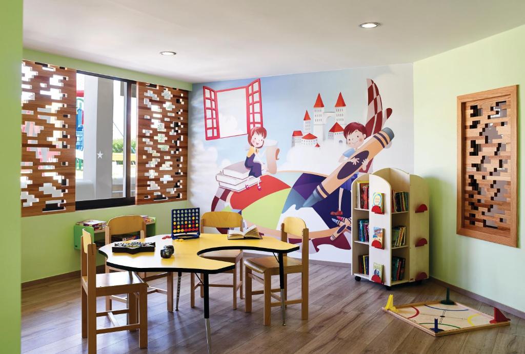Krāsaina bērnu istaba ar galdu un krēsliem, uz sienas ir bērnu ilustrācija ar grāmatām un zīmuli. Istabā ir arī plaukti ar grāmatām un spēlēm, radot radošu un mācību vidi.