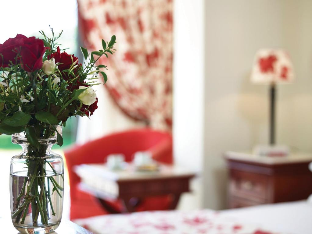 Sarkano rožu pušķis caurspīdīgā vāzē rotā elegantu istabu, kurā redzama sarkana atpūtas krēsls un lampiņa ar ziedu motīvu.