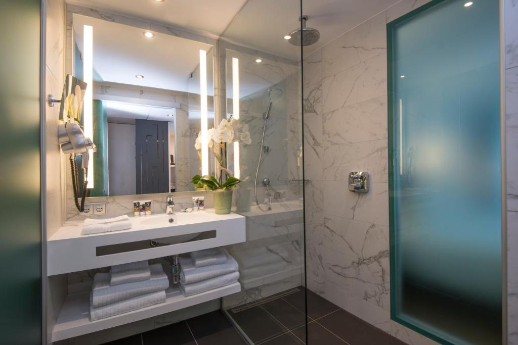 Mūsdienīgs vannas istabas interjers ar marmora sienām, stikla dušas kabīni un modernu izlietni. Telpā ir eleganta apgaismojuma sistēma un dekoratīvas orhidejas.