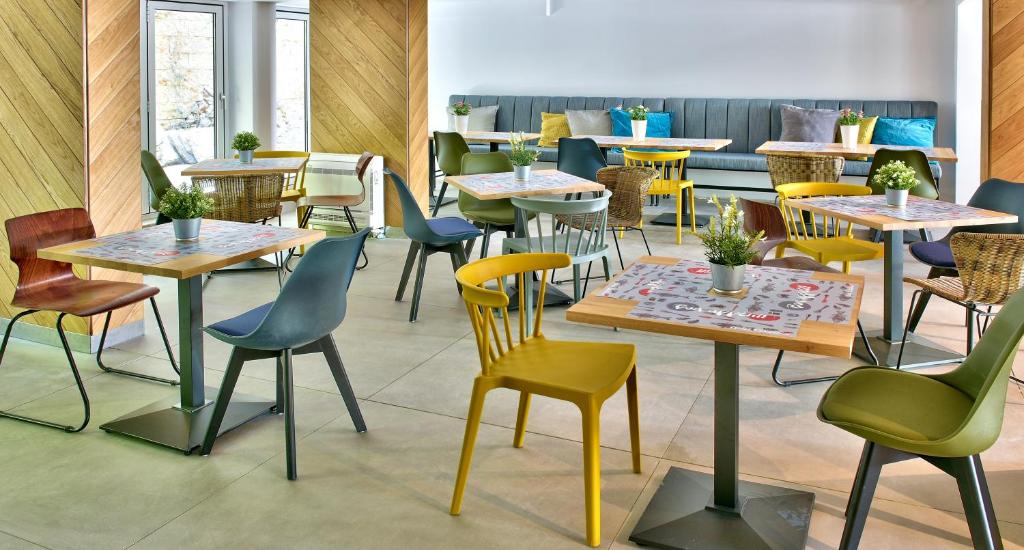 Mūsdienīgs kafejnīcas interjers ar dažādu krāsu un dizaina galdiem un krēsliem, kā arī dekoratīvām puķu podiņām uz galdiem. Telpa ir gaiša un aicinoša, radot patīkamu atmosfēru.