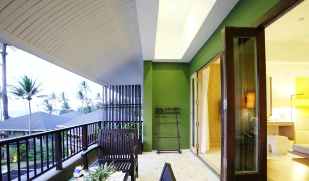 Balkons ar zaļām sienām, koka mēbelēm un skatu uz palmu dārzu. Telpa ir gaiša un moderna, radot relaksējošu atmosfēru.