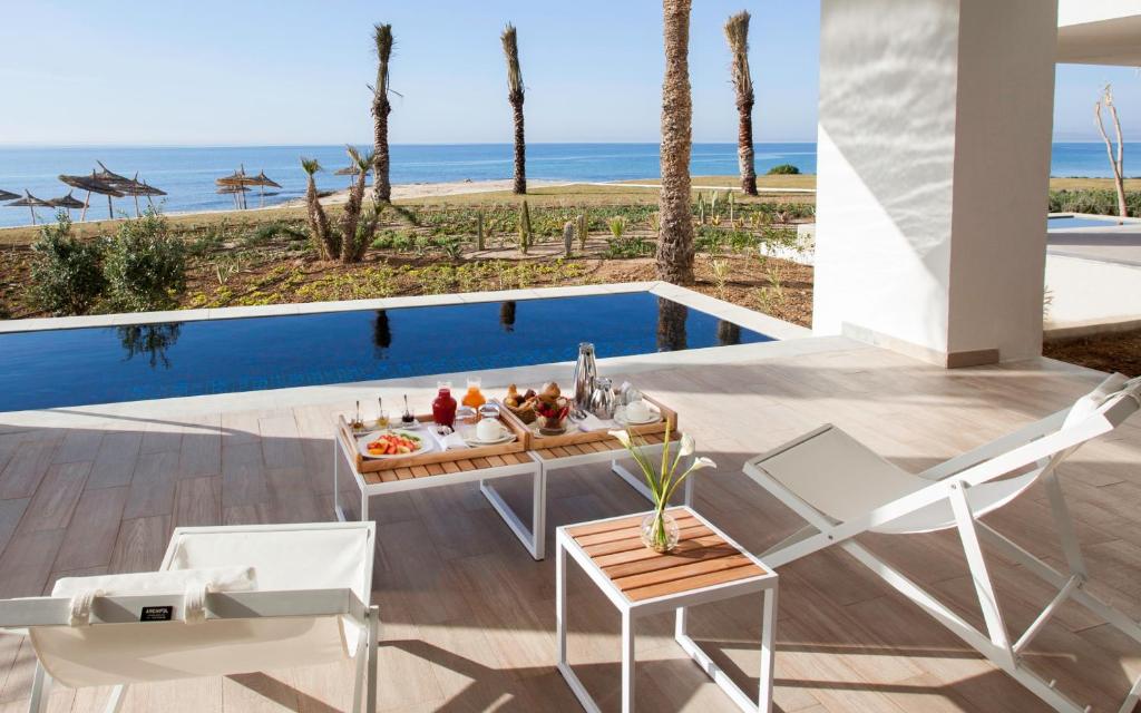 Atjaunojoša pludmales skats no terases ar baseinu, kur uz galda ir brokastu uzkodas un dzērieni. Apkārt aug palmām un pludmalei.