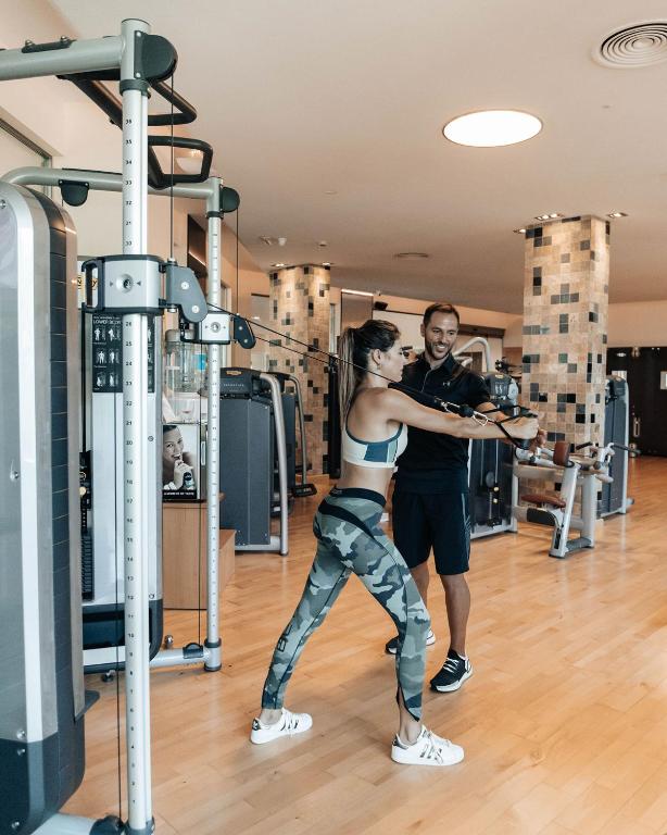 Sieviete trenējas sporta zālē ar trenera palīdzību, izmantojot stieņa iekārtu. Aiz viņas redzami sporta aprīkojumi un moderna vide.