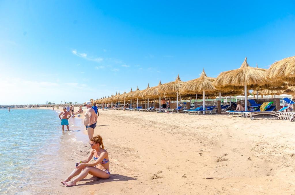 Pludmales attēls ar saulē sildošiem apmeklētājiem, kas atpūšas pie ūdens un zem šūpuļtīkliem. Skats uz skaistu jūru un saulainu debesi.