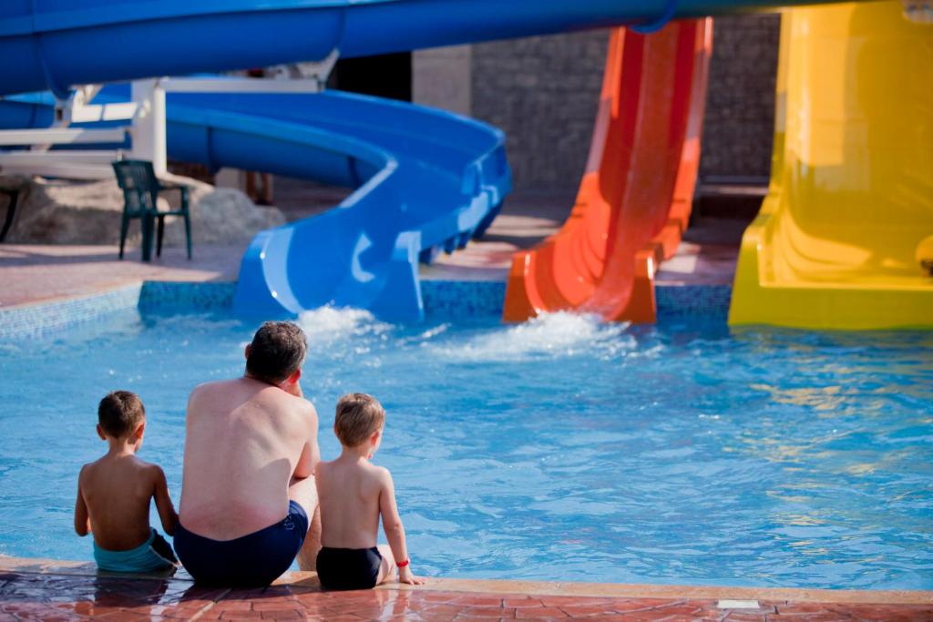 Divi bērni un pieaugušais sēž pie peldbaseina, vērojot ūdens slaidus ar spilgtām krāsām. Ūdens izšļakstās no slaidu gala, radot jautras brīvdienu noskaņas.