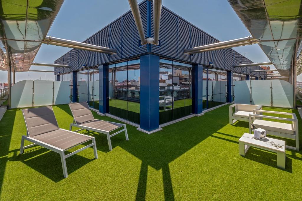 Terases zona ar mākslīgo zālieni, kur atrodas divi sauļošanās krēsli un balta dīvāna komplekts, kas piedāvā relaksējošu atmosfēru. Caur stikla sienām redzams skats uz apkārtējo pilsētu.