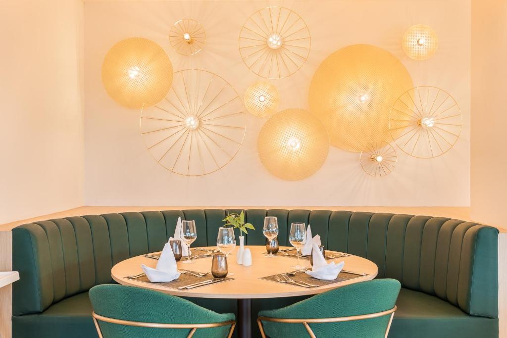 Eleganta restorāna iekārtojums ar zaļu dīvānu un dekoratīvām gaismām uz sienas, radot siltu atmosfēru. Galdiņš ir sakārtots ar galda piederumiem un ziedu kompozīciju.