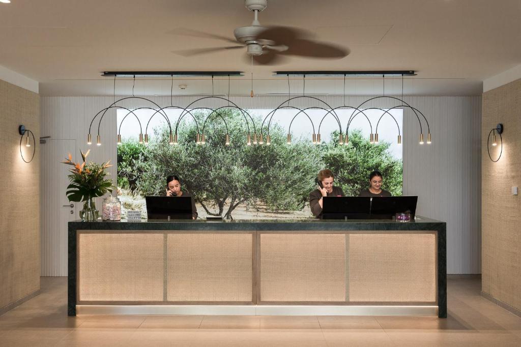 Mūsdienīgs viesnīcas reģistratūras galds ar darbiniekiem, kas apkalpo viesus. Aiz galda redzams dārza skats ar olīvu kokiem, radot patīkamu atmosfēru.