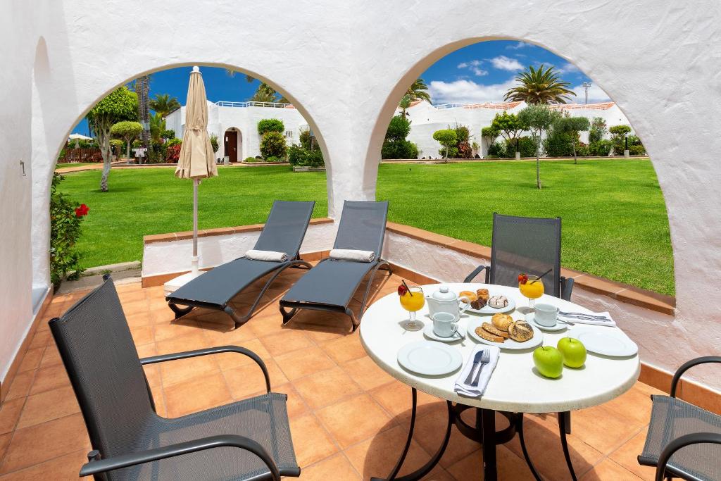 Mūsdienīgs terases skats ar galdu, uz kura ir brokastu ēdieni un dzērieni, kā arī divas sauļošanās krēsli. Fonā redzams zaļš dārzs ar palmām un ziediem.