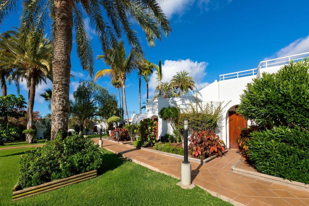 Vasaras māja ar zaļu dārzu, palmu kokiem un skaidru zilu debesi, radot mierīgu un relaksējošu atmosfēru.