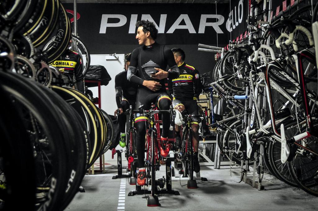 Velosipēdisti trenējas slēgtā telpā ar daudzām velosipēdu riepām un aprīkojumu, sagatavojoties sacensībām. Fons ir melns ar Pinar logo.