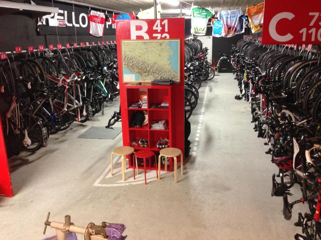 Velosipēdu novietne ar daudzām velosipēdu rāmjiem, kas piestiprināti pie sienām, un sarkanu stendu, kurā ir kartes un apavi. Gaisotne ir organizēta un sportiska.