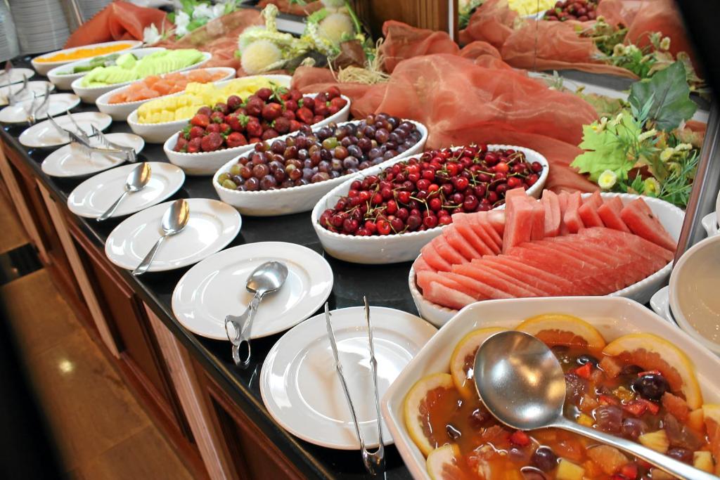 Dažādas svaigas ogas un augļi, ieskaitot arbūzus, zemenes, vīnogas un ananāsus, izkārtoti uz galda ar baltiem šķīvjiem un karotēm.