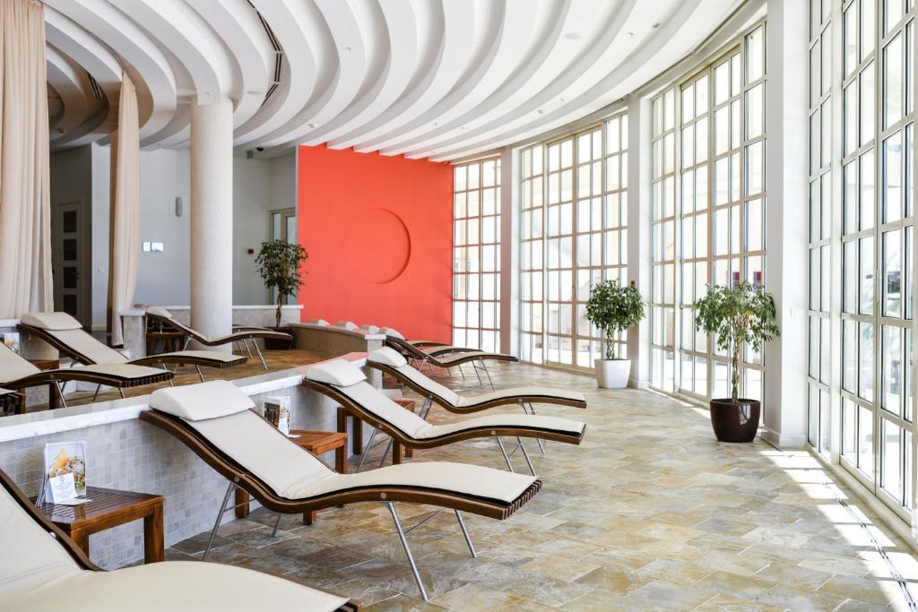 Mūsdienīgs spa telpas interjers ar relaksācijas krēsliem un plašiem logiem, kas nodrošina dabīgu apgaismojumu. Telpa ir veidota ar elegantu dizainu un augu dekorācijām, radot mierīgu atmosfēru.