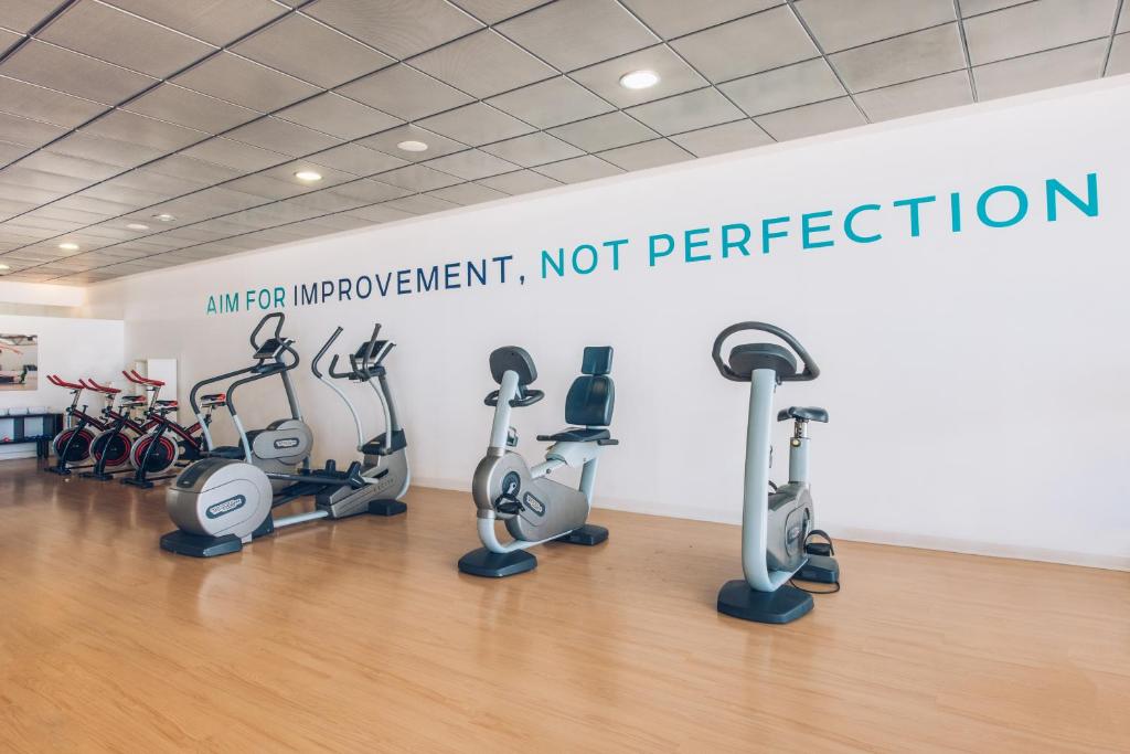 Mūsdienīgs sporta zāles interjers ar dažādām trenažieru iekārtām un motivējošu uzrakstu "Mērķis ir uzlabojums, ne perfekcija".