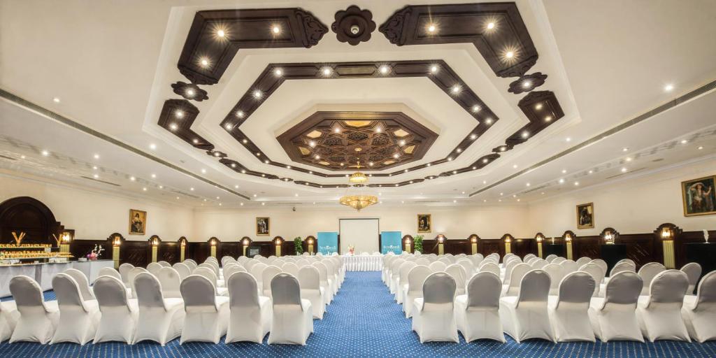 Elegants konferenču zāles interjers ar baltiem krēsliem un greznu griestu dizainu. Telpa ir gatava pasākumam vai sanāksmei, piedāvājot plašu un komfortablu vidi.